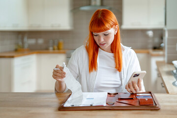 A young woman with vibrant red hair reviews documents on a kitchen table while using her phone in a modern home setting