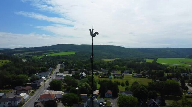 Aerial drone view of the rooster‑shaped weather vane and ornate wrought iron cross crowning the church steeple spire in the rural village of Saint‑Angele‑de‑Merici in Quebec, Canada