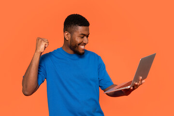 A joyful man celebrates a victorious moment while using a laptop against a vibrant orange backdrop