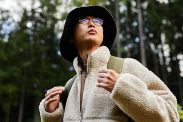 Portrait of young Asian man backpacking in forest and looking up with focus on eyeglasses and comfy...