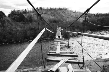A girl walks across an old wooden suspension bridge over a river in an autumn forest. The scene emphasizes the atmosphere of adventure and solitude in the wild.