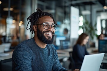 Obraz premium A joyful young professional wearing glasses and a sweater, sitting in a modern office environment, and working on a laptop with a bright smile.