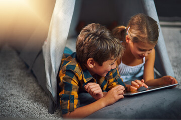 Tablet, rest and kids in tent in home playing online educational game for learning. Relax, digital technology and children siblings watching video for entertainment in blanket fort for fun at house.