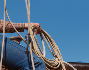 Close-up photo of a cowboy's hand holding a coiled lasso rope while sitting on horseback, wearing a...