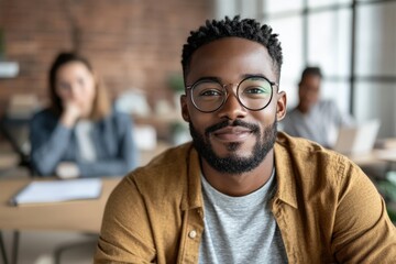 Fototapeta premium A confident young man with glasses sits in a bright office, his demeanor friendly and approachable, reflecting a modern professional environment of collaboration and openness.