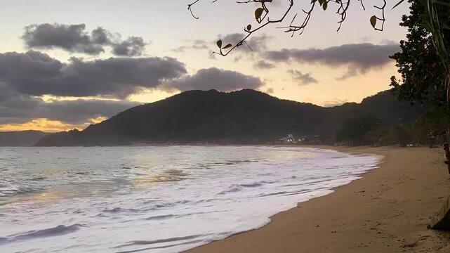 Time-lapse final de tarde com o mar forte, a praia vazia e muitas nuvens passando atr&aacute;s da montanha ao fundo. Dois cachorros passam, aproveitando a tranquilidade.
