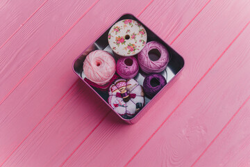 A top view of a tin box containing vibrant yarn and sewing items, placed on a pink wooden surface....