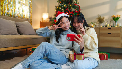 Two friends in holiday headwear smiling as they look at a phone, sitting beside a decorated...