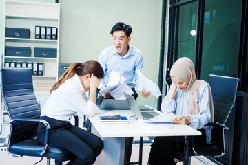 Fototapeta premium A Muslim businesswoman sits with an Asian man and woman, all middle-aged, at a desk with a laptop, appearing stressed as they discuss challenging business matters in a corporate setting