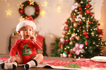 Cute little boy wearing Santa hat sitting on table, playing with toys on Christmas day and Christmas tree background.