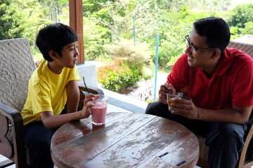 A father and son enjoy quality time together while sitting outdoors at wooden table.Relaxing with refreshing drinks,the serene greenery around them adds to the peaceful ambiance of their conversation