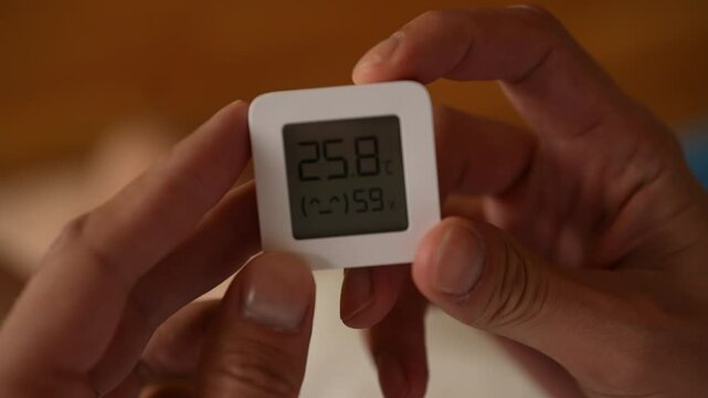 Male hands holding small digital hygrometer and thermometer, displaying temperature and humidity, monitoring indoor climate conditions, closeup, unrecognizable person. Shooting in slow motion.