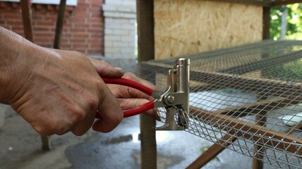 male hands holding red handled construction pliers and clamping metal mesh to make a rim of the...