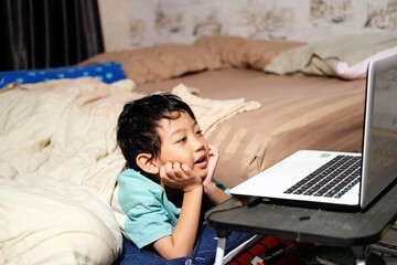 A young boy lying on his bed, watching something on a laptop with a look of fascination. He appears engaged and comfortable in a cozy, home setting, enjoying screen time.