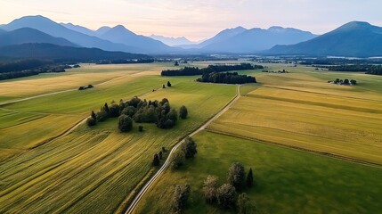 Fototapeta premium Aerial view of expansive agricultural fields with a winding path, surrounded by distant mountains under a clear sky at dusk.