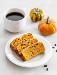 Slices of pumpkin sweet bread or cake with icing on a plate on a light background with decorative pumpkin and a cup of coffee.