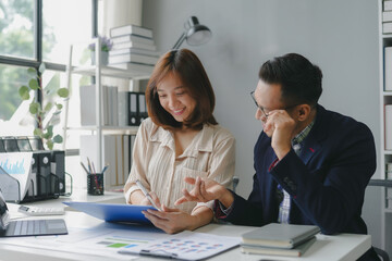 Two asian coworkers are discussing and working together using a digital tablet and looking at paperwork with graphs and charts