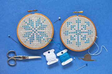 Top view on two hand embroidered cross stitch snowflakes in hoops on blue background, accessories for cross stitch. Winter crafts, Christmas gifts concept. Cross stitches as hobby. Flat lay, close-up