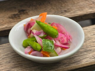 A small white bowl containing a colorful mixture of pickled vegetables, including green chilies, sliced carrots, and red onions, placed on a wooden surface