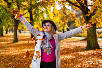 Portrait of happy middle-aged woman wearing gray coat and scarf, pink sweater, black brimmed hat throwing golden leaves against alley with autumn trees in background in city park on sunny day. Energy 