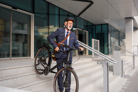 Smiling elderly businessman carrying bicycle and moving down on staircase while leaving from office