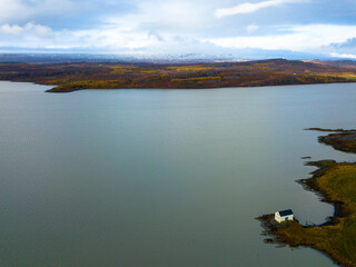Small white house reflecting on the calm water of lake Lagarflj?t in Egilssta?ir, Iceland, on a cloudy day with snowy mountains in the background