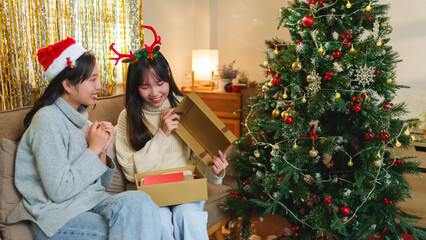 Two friends opening a gift box by the Christmas tree, sharing a joyful moment in a cozy holiday...