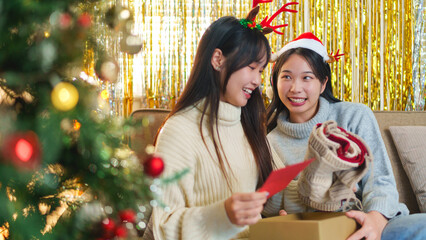 Two friends sitting on a couch, joyfully unwrapping a holiday gift together with Christmas decorations around them, wearing reindeer antler headbands.