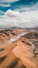 Fototapeta premium Aerial view of vast, rolling sand dunes and a small town nestled in the valley below, under a blue sky with fluffy white clouds.