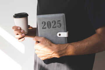 male hand holding a diary with the text 2025 and paper cup with hot drink on the background of a...