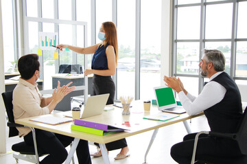 Fototapeta premium Asian businesswoman wearing mask and presenting chart work on a glass board to Caucasian manager and co-workers in an office conference room for project meeting