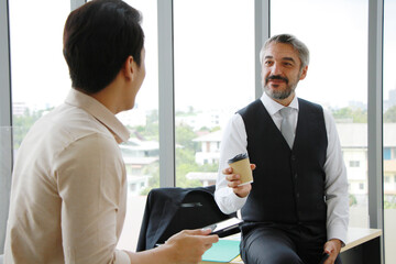 Smiling elderly senior Caucasian manager drinking coffee and talking with asian young businessman for brainstorming for project job together in office