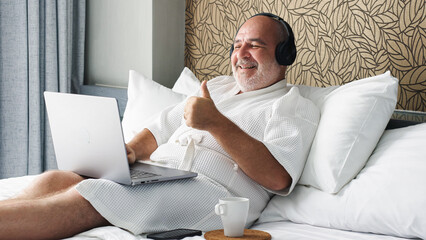 A man on vacation attends a video conference in a bathrobe, sitting on the bed with headphones and a laptop. A casual yet professional scene, blending relaxation and remote work.