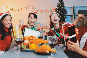 Group of young Asian man and women as friends having fun at a New Year's celebration, holding gift boxes standing by Christmas tree decoration, midnight countdown Party at home with holiday season.
