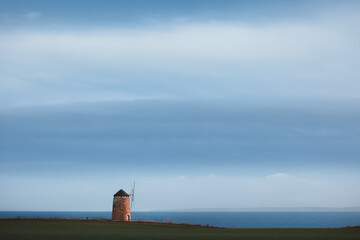 A lone St Monans Windmill stands serenely on the horizon under a moody blue sky, overlooking the...