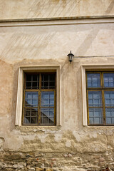 Two rectangular windows with bars. Windows of an old castle building. Decorative lamp on the wall above the windows. Old decaying facade. Windows, glass, lamp, fallen facade, rough, broken