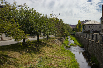 photo of a small stream with a concrete bed. Tree-lined avenue along the stream.  The main street of a small village with a stream running through it. River, grass, trees, houses, sky.