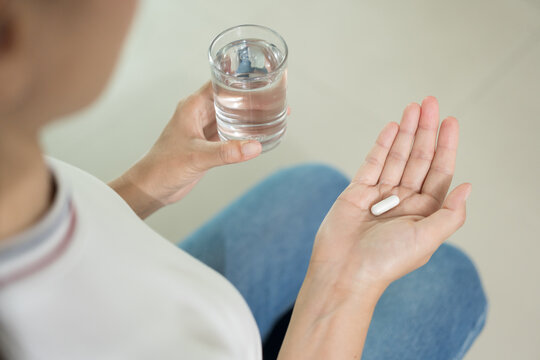 Closeup woman holding pills capsule time to take medications. Sick female taking medicine drinking with water glass sit on sofa at home. Healthcare concept.