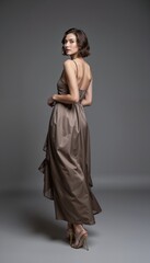 Fototapeta premium Elegant young woman in taupe gown posing with back turned against gray background 
