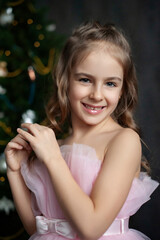 A young girl in a pink dress stands in front of a beautifully decorated Christmas tree. The photo is captured in low-key lighting, which creates a dramatic, moody atmosphere. magic Christmas.