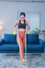 Fototapeta premium Concentrated young sportswoman is performing leg stretching exercises using a resistance band while working out in her living room