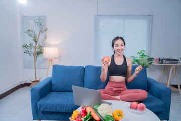 Young woman nutritionist showing healthy food, holding an apple and broccoli, sitting on a blue couch with a laptop and fresh vegetables and fruits on the table in her living room