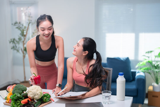 Two asian nutritionists are smiling and creating a personalized balanced diet for their customer, using fresh vegetables and taking notes on a clipboard
