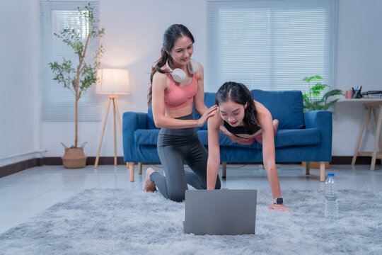 Two young asian women are doing fitness exercises at home, following an online class on a laptop, one woman is doing push-ups while the other one is controlling her posture