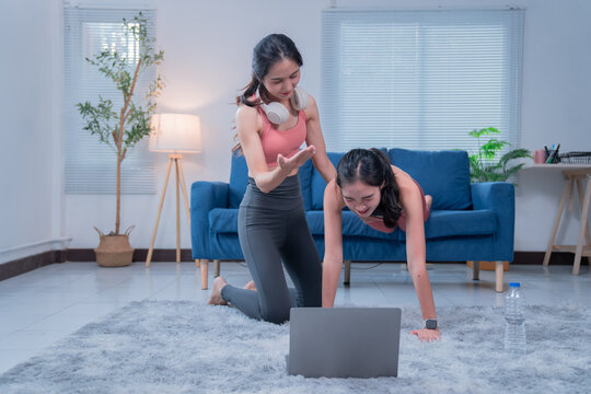 Two young asian women wearing sportswear are doing a home workout following instructions on a laptop, with one woman performing push-ups while the other gives instructions