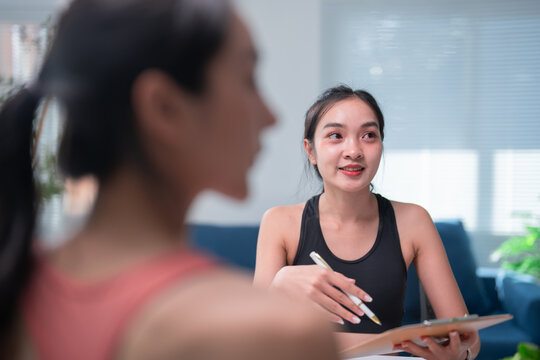 Asian female trainer in sportswear explains a workout plan to her client in a living room filled with natural light, emphasizing fitness and health - Powered by Adobe