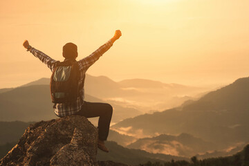 The traveler with a backpack raises his hand proudly for conquering a mountain peak. A hiker sits with his arms outstretched to convey success and freedom atop a misty high mountain