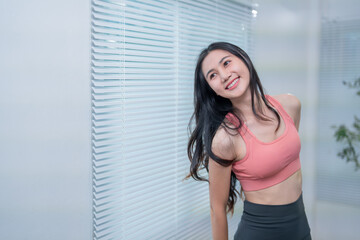 Asian fitness woman in sportswear smiles and relaxes at home after a workout, basking in natural light by a window. Her content and healthy lifestyle exude motivation and inspiration