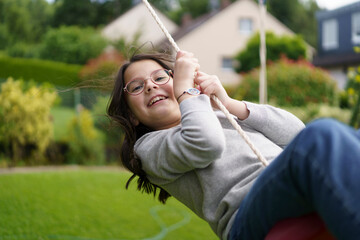 A cute little cheerful girl in glasses and a sweater is riding a swing in the backyard of her...
