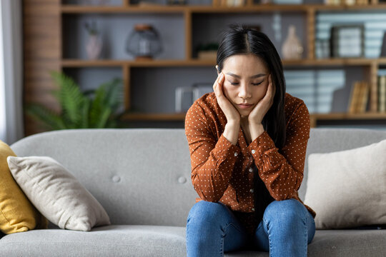 Asian woman sitting on sofa in cozy home, looking thoughtful. Casual attire suggests relaxed setting. Neutral expression hints at contemplation, reflection in comfortable environment.